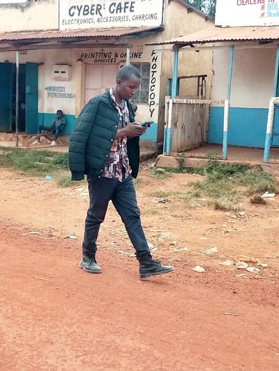 The image shows a man walking down a dirt road in Kivaywa, Kenya, located at latitude 0.562 and longitude 34.805. The man is wearing a dark green jacket, a multi-colored shirt, dark blue pants, and black shoes. He is looking at a mobile phone in his right hand. The road is red, and there are buildings on either side. One building has a sign that reads "Cyber Cafe, Electronics, Accessories, Charging." Another building has a sign that reads "Printing & Online Services."  There is also a sign that reads "Photocopy." The man is walking towards the right side of the image.  There is a small amount of vegetation on the sides of the road.  There is a man sitting on a bench behind the person in the foreground. The man is wearing a dark blue jacket.  The background is out of focus.