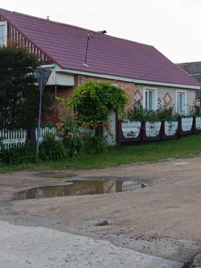 The image depicts a rural scene in Smaliavitski Sel'ski Savet, Belarus. A modest brick and white painted house with a burgundy metal roof dominates the background. The house has several windows and is partially obscured by lush green foliage. In front of the house, there is a white picket fence interspersed with decorative brown sections, and the fence is further softened by flowering plants. A dirt road with puddles of water is in the foreground, reflecting the fence and greenery. A triangular signpost stands to the left of the house. The overall impression is of a quiet, well-maintained rural dwelling surrounded by nature.