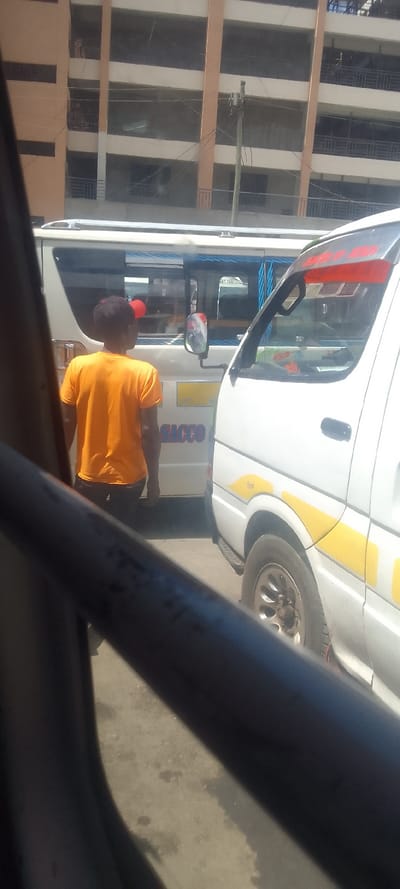 The image depicts an urban street scene in Naivasha, Kenya, viewed from within another vehicle. In the midground, a male individual stands facing away from the viewer, positioned between two white public service vans (matatus). He wears an orange short-sleeved t-shirt, dark trousers, and a red baseball cap. The matatu to the viewer's right is partially visible, showing its white front quarter, a black side mirror, a red sun visor above the windshield, and yellow curved decals along the side. A silver multi-spoke wheel is visible below the vehicle's body. The matatu to the viewer's left is also partially visible, displaying the word "SACCO" in blue lettering on its side and a blue rope-like trim along its roofline. Both vehicles appear to be parked or stationary on a paved, slightly stained ground surface. In the background, a multi-story building with light brown vertical pillars and darker horizontal recessed sections, possibly containing windows or balconies with dark railings, dominates the skyline. A wooden utility pole with multiple overhead wires stands in front of this building. A dark, out-of-focus metal bar or frame from the immediate foreground partially obstructs the lower portion of the image. The scene is illuminated by bright daylight.