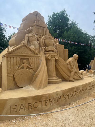 This media file showcases a large, intricate sand sculpture, likely an exhibit from a festival or event. The sculpture is primarily made of sand and depicts a complex scene with human figures and architectural elements.
The main subjects include at least two human figures rendered in sand. One figure appears to be seated, possibly playing an instrument or holding something, and is wearing a helmet and clothing reminiscent of an aviator or historical figure. Another figure is depicted in a more stylized, almost bas-relief form within an architectural detail, appearing to be a dancer or performer in a flowing gown. Architectural elements such as columns with Ionic volutes and layered structures resembling buildings are incorporated into the sculpture, suggesting a grand or historical theme.
The setting appears to be outdoors, with trees visible in the background and a cloudy sky overhead, suggesting it might be daytime. Small flags, possibly representing different nations or organizations, are strung up behind the sculpture, hinting at an international event or exhibition. A rough rope barrier is visible in the foreground, indicating the sculpture is on display and protected from direct public access.
A prominent detail is the carved text on a curved surface of the sculpture. The text is in Cyrillic script and appears to read "НАВСТРЕЧУ КУЛЬТУРНОЙ СТОЛИЦЫ" which translates to "Towards the cultural capital." This text provides a significant clue to the theme or purpose of the sculpture, likely celebrating or anticipating a city being designated a cultural capital. The sand itself is a warm, light brown color. The overall impression is one of artistry and community engagement, with the sculpture serving as a focal point for a public event.