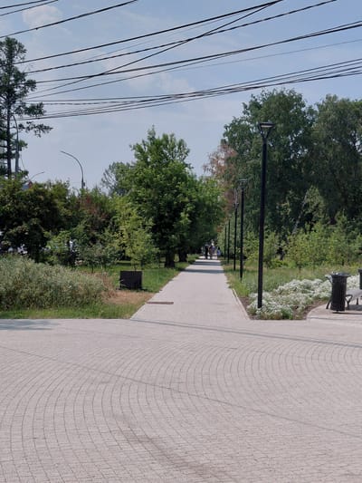 The image depicts a paved pathway in a park setting, likely in the city of Novosibirsk, Russia, given the common architectural and landscaping style. The pathway is constructed of light-colored paving stones laid in a curved pattern, leading into the distance. It is bordered by green grass and lush trees on both sides. In the foreground, the pathway curves to the right, revealing more of the paving stone pattern. Several modern lampposts are visible along the pathway, suggesting it's a public space designed for evening use. In the background, a few people are walking along the path, their forms somewhat indistinct due to the distance. The sky above is a pale blue with scattered white clouds. A prominent feature is the network of overhead power lines, a common sight in urban environments. Further along the path, to the right, there is a park bench and a trash receptacle. The overall scene conveys a sense of a well-maintained urban park, inviting for leisure and recreation.