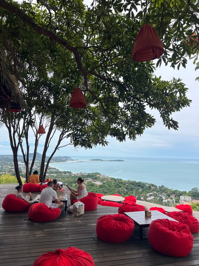In Baan Chaweng Noi, Thailand, a scenic open-air restaurant offers a panoramic view of the turquoise sea and a lush green hillside. The foreground is dominated by a wooden deck furnished with numerous vibrant red beanbag chairs and low wooden tables. Several groups of people are seated, some engaged in conversation, others reading, while enjoying the stunning vista. A few red, conical lamps hang from the branches of large trees that provide ample shade over the seating area. The background reveals the coastline dotted with buildings and the vast expanse of the ocean meeting the horizon under a slightly overcast sky.