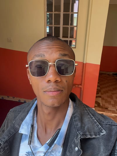 A medium shot depicts a dark-skinned man with short hair and sunglasses, wearing a patterned shirt and a dark denim jacket. He also has wired headphones around his neck. The background reveals a yellow and red wall, and a window with multiple panes. The location is likely in Nyeri, Kenya, as specified in the prompt.