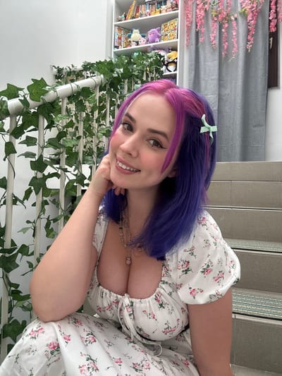 A person with pink and purple hair is smiling at the camera. They are wearing a white floral print dress and leaning against a white railing covered in fake ivy. Behind them is a bookshelf filled with various items and a gray curtain. In the background, there are stairs with silver accents and a gray wall. The location is possibly indoor, somewhere in the city of Zagreb, Croatia.
