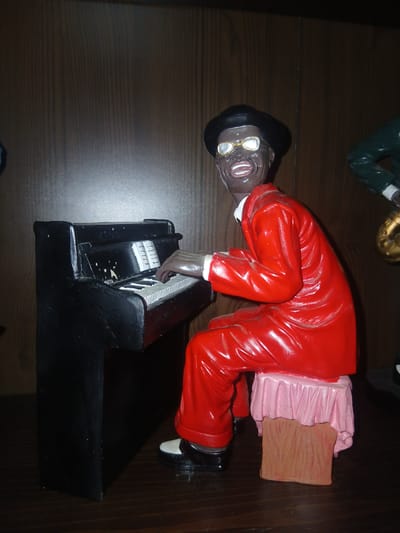 This is a close-up, eye-level shot of a figurine of a Black man playing a piano. The figurine is a dark brown color with wide, grinning teeth and bright white eyes. He is wearing round, gold-rimmed glasses and a black hat. His outfit consists of a bright red suit and white shirt. The piano is black and appears to be a toy or decorative piece. The figurine is seated on a stool covered with a pink cloth with ruffles, which is resting on a brown, wood-grain block. The background is a dark wooden surface. The figurine is positioned to the left of the frame, with the piano in front of it. The right side of the image shows a portion of another figurine, which appears to be wearing a green jacket. The lighting suggests the object is illuminated from the left, casting a shadow to the right. The image lacks any discernible details that would indicate a location in Navaluenga, Spain.