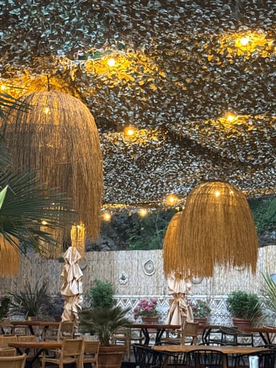 This media file captures an outdoor setting with a focus on decorative elements and dining furniture. The scene appears to be set at dusk or evening, as evidenced by the string lights illuminating the area. The background features a textured wall covered in what looks like foliage or a patterned textile, interspersed with glowing yellow lights.
Prominently featured are two large, dome-shaped objects made of straw-like material, possibly lampshades or decorative features, each with internal lighting casting a warm glow. Below these structures, draped fabric adds to the ambiance. In the background, a rustic wall with geometric patterns is visible, along with potted plants and greenery, suggesting an outdoor dining or lounge area.
To the right of the frame, a collection of wooden tables and chairs, some with woven seats, are arranged. This arrangement indicates a dining or seating area. The overall impression is one of a relaxed, perhaps restaurant or cafe setting, in Kamenovo, Montenegro, designed to create a cozy and inviting atmosphere. There is no visible text in the image.
