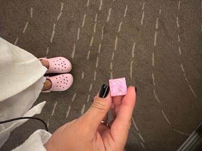 The image shows a close-up of a hand holding a small, pink, square object. The object appears to be some type of candy or trinket with a textured surface, possibly featuring a raised image or pattern. The hand has dark, black painted fingernails and a ring on the ring finger. In the background, a person's feet are visible wearing pink Crocs, partially covered by a white cloth. The flooring appears to be a textured carpet with a repeating pattern of light-colored lines. The scene is indoors, likely a domestic setting. The image does not provide any information about the location in the city of Žunovnica, Bosnia and Herzegovina.