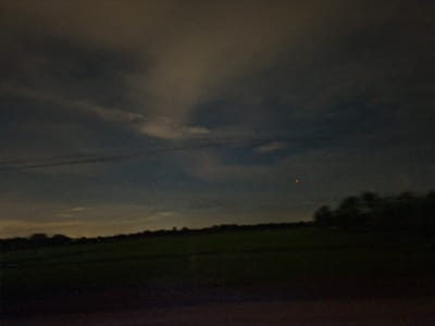 The media file depicts a dusky landscape, likely captured during twilight or at night, given the low light conditions and the presence of a small, orange, circular object in the sky, possibly a light or a distant celestial body. The scene is dominated by a dark, cloudy sky. Below the sky, a silhouetted treeline marks the horizon, behind which lies a dimly lit, open field, likely agricultural. The foreground is a dark, blurred area, suggesting the image was taken from a moving vehicle or a low vantage point. The overall impression is one of quietude and perhaps a touch of mystery due to the unexplained light in the sky. There is no visible text within the media file. The location context is Dong Nai City, Vietnam.
