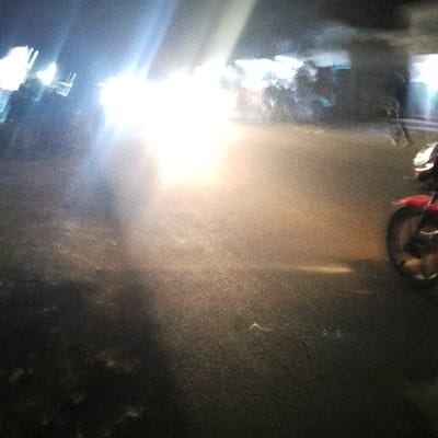 A blurry, low-light image features a street scene at night. Bright headlights illuminate the road, creating a hazy glare. A red motorcycle is visible on the right, while other vehicles are indistinct in the background. The asphalt road appears wet and reflective. Overall, the photo conveys a sense of motion and poor visibility.