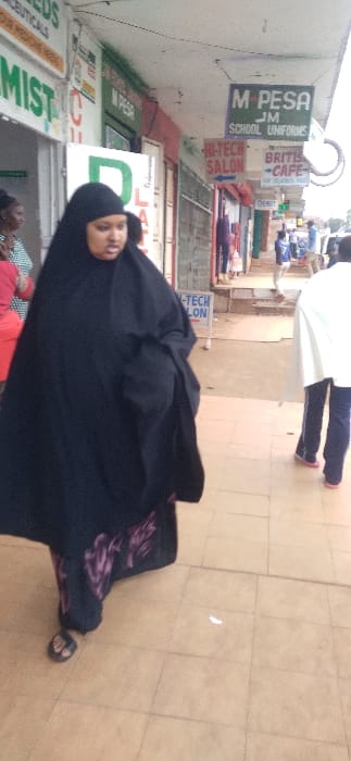 A daytime street scene in Nyeri, Kenya, depicts a woman walking from left to right on a light brown tiled sidewalk. She is wearing a black abaya, which covers a garment with a visible purple floral pattern, and black open-toed sandals. Her face is exposed, with light brown skin and dark hair. To her immediate left, partially obscured, is another individual in a light-colored striped top.

The background features a row of commercial storefronts. Visible signage includes a partial green "PHARMIST" sign, a "M-PESA" sign over a doorway, a larger overhead sign for "M-PESA JM SCHOOL UNIFORMS," a red "HI-TECH SALON" sign with a left-pointing arrow, a "BRITISH CAFE" sign advertising "FOR DELICIOUS FOOD," and a small green "CHEMIST" sign with a right-pointing arrow. Buildings display red and green accents. Further down the street to the right, a person in a white garment, dark trousers, and pink shoes walks away from the camera. The environment beyond the tiled sidewalk shows unpaved ground leading to a distant street with additional indistinct pedestrian figures. The sky is overcast, indicating diffused daylight.