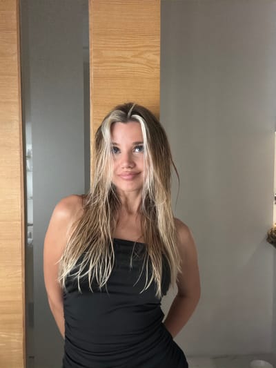 The image shows a medium shot of a young woman with blonde and brown hair, styled in loose waves. She is wearing a black strapless dress and has her arms behind her back. Her expression is neutral. The background appears to be an indoor setting, possibly a hotel room or bathroom, with a wall and a wooden partition. There are no visible indicators to suggest the location is in Thep Krasatti, Thailand.