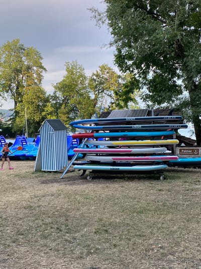 The image depicts an outdoor scene in Milvignes, Switzerland, likely at a watersports rental facility. In the foreground, a dry, grassy area is visible, with a few scattered leaves. To the left, a young person wearing a life vest and pink shoes is walking and holding an orange paddle. Behind them are rows of blue paddle boats. In the center of the image is a small, striped blue and white hut. To the right, a rack holds multiple stand-up paddleboards of various colors, including dark blue, teal, yellow, pink, and light blue. Some boards have visible branding such as "LOKAHI," "AQUADISIGN," "MALIBU 10'6 X 32," and "OCEAN PACIFIC 10'6 X32". A sign reading "Palmar Embarkation point" with a boat symbol is also visible. In the background, tall green trees with dense foliage form a backdrop, and beyond them, a body of water and hills are faintly visible under a cloudy sky. The scene suggests a recreational area by a lake or river, possibly during late afternoon or early evening given the lighting.