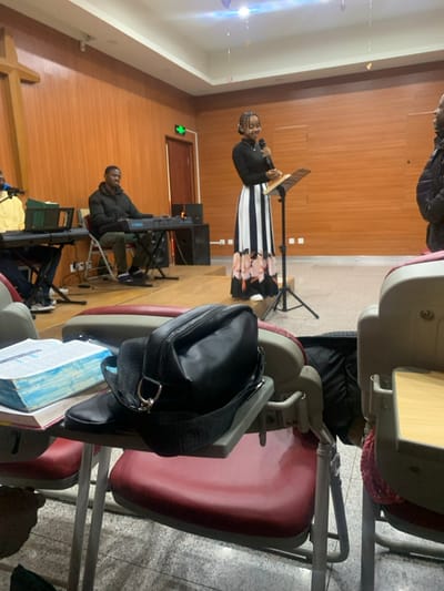 This image depicts an indoor gathering within a hall in Shenyang City, China. The setting features wood-paneled walls, a light-colored floor, and overhead lighting. A raised platform serves as a stage in the background.

On the stage, a Black woman stands center-right, holding a microphone in her right hand and addressing an unseen audience. She wears a black long-sleeved top and a long skirt with black and white vertical stripes transitioning to a floral pattern. To her left, two Black men are seated behind electronic keyboards. The man closer to the woman wears a dark puffer jacket and operates a Yamaha keyboard. Another man in a yellow shirt is partially visible on the far left, behind a second keyboard. A large wooden cross is partially visible on the far left wall, indicating a religious context. Black speaker cabinets are positioned on the stage floor.

In the foreground, red chairs with integrated folding wooden desks are arranged, facing the stage. On one of these desks, a black shoulder bag with a silver D-ring buckle rests on top of a stack of books, one of which has a prominent light blue cover. Small, light-colored decorative elements are suspended from the ceiling. A green "Exit" sign is visible above a dark door on the back wall. No specific landmarks or identifying features are discernible to pinpoint a precise location within Shenyang City beyond the characteristics of a communal hall or religious institution.