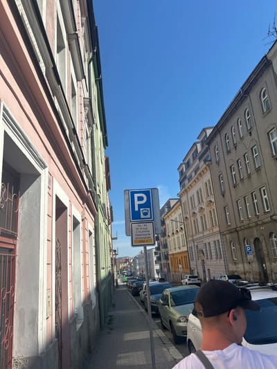 This is a street-level, slightly upward-angled, medium shot of a narrow street in Pilsen, Czechia, on a sunny day. The perspective is from the sidewalk on the left side of the street, looking down the road. On the left are the facades of several old buildings with pastel-colored exteriors, including pink and green. These buildings feature multiple stories with windows and decorative architectural elements. A blue and white parking sign with text in Czech is prominently displayed on the right side of the sidewalk. Further down the street, on the right, are more multi-story buildings with grey and cream-colored facades. Parked cars line the right side of the street. In the foreground, a person wearing a black baseball cap and a white t-shirt is partially visible from the back, facing away from the camera and towards the street. The sky is a clear, bright blue. The overall scene depicts a typical urban streetscape with residential and commercial buildings.