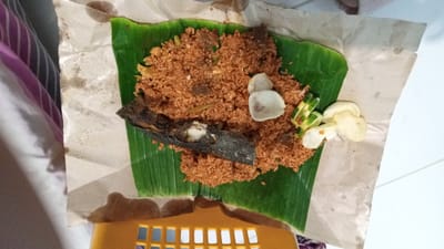 This media file displays a close-up overhead shot of a meal served on a banana leaf, placed on crumpled brown paper. The meal appears to be a portion of fried rice, a dark-skinned fried fish, some sliced cucumber, and what look like crackers or crisps. The fried rice is a reddish-brown color and is mixed with small pieces of meat or vegetables. The fish is positioned horizontally across the rice. The banana leaf is vibrant green and forms a natural serving platter. The setting is simple, possibly a casual dining environment or street food stall in Buketteukuh, Indonesia. There are no people visible, and the focus is entirely on the food. The lighting suggests an indoor setting or perhaps an overcast day. The overall presentation is rustic and informal. No text is visible in the image.