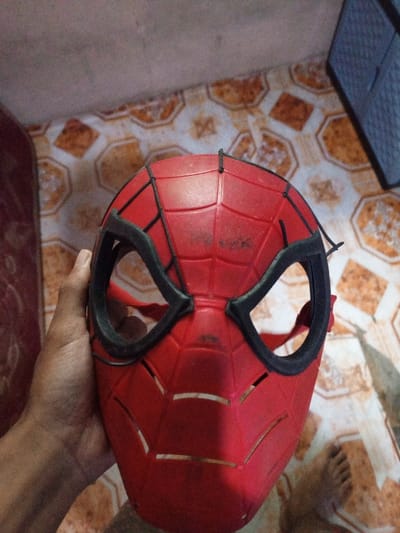 A person's hand holds a red Spiderman mask with black web designs and eye frames. The mask is visibly worn, showing some smudges and signs of use. It has horizontal slits below the eye area. The background includes a tiled floor with an orange and brown pattern, part of a bed with red fabric, and a grey plastic container, creating a slightly cluttered domestic setting. A foot is also visible in the background.
