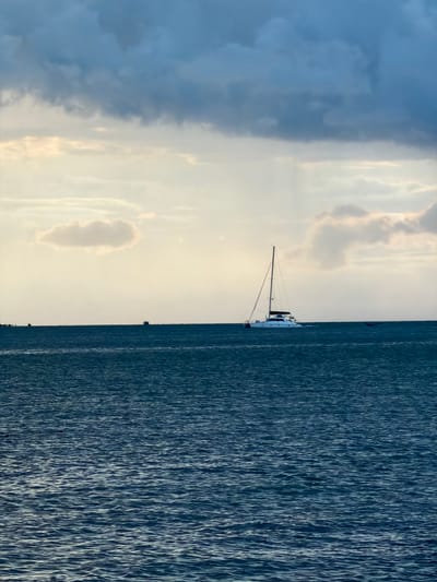 This is a rotated image of a catamaran sailboat on the water, with a cloudy sky. The catamaran is white with a tall mast and appears to be moving. The sea is dark blue and choppy, with whitecaps visible. The sky is a mix of dark and light clouds, with some areas of sunlight breaking through. The image seems to be taken during the day, likely in the late afternoon or early evening, given the soft light. There are no people or text clearly visible in the image. The scene evokes a sense of peacefulness and the vastness of the ocean. The location context suggests it was taken near Baan Bo Phut, Thailand.