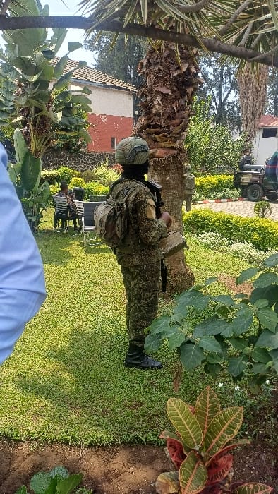 The image displays an outdoor scene within a well-maintained garden in Goma, Democratic Republic of the Congo.

In the central foreground, a person in full military-style digital camouflage uniform stands with their back to the viewer, positioned slightly right of center. This individual wears a camouflage helmet, a dark balaclava covering the lower face and neck, black boots, and a light-colored camouflage backpack. A dark object, consistent with a firearm, is held in the right hand, while a large, flat, rectangular, olive-green box, possibly an ammunition container, is held in the left hand, resting at hip level.

Immediately in front of the uniformed person is the textured, brown trunk of a large palm tree, its fronds extending out of frame upwards. The ground is predominantly covered in short, manicured green grass. In the lower right foreground, a plant with variegated red and green leaves is visible. To the far left foreground, the partial shoulder and arm of an individual in a light blue collared shirt are present, indicating an observer's perspective.

In the midground, to the left of the uniformed person, two individuals in dark clothing are seated at a dark-colored table with two chairs on the grass, facing each other. Behind them, a low stone wall is visible. To the right of the uniformed person, a paved area featuring a red and white patterned curb is present. A dark-colored utility vehicle, likely an SUV with a rear-mounted spare tire, is partially visible on this paved area.

The background includes a two-story building to the left, featuring a reddish-brown tiled roof, a light-colored upper facade, and a reddish-brown lower section with a small rectangular window. Extensive green foliage and numerous trees, including another palm tree trunk, are visible across the background. A clear blue sky is visible above the structures and trees.