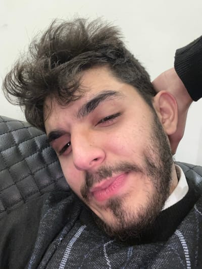 A close-up image depicts a young adult male in an indoor environment within the city of Rukn ad-Din Municipality, Syria. The subject has dark, unkempt curly hair, dark eyebrows, a mustache, and a full beard. His eyes are partially closed, and his lips are closed and slightly upturned at the corners. He is wearing a dark-colored, possibly knit sweater with a subtle geometric pattern, over a white collared shirt. The man's head is resting against a dark, quilted, cushioned surface, consistent with an upholstered chair or sofa. A plain, light-colored wall forms the background in the upper right. A hand, partially obscured by a dark sleeve, is positioned behind the subject's right ear.