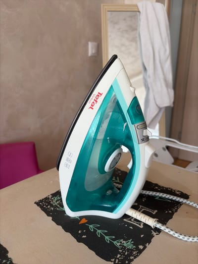 The image shows a close-up of a Tefal Virtuo steam iron, predominantly white with teal accents. The iron is positioned diagonally on a textured surface, likely a piece of furniture, with a patterned cushion visible to the right. A white and grey patterned power cord extends from the back of the iron. The background is softly blurred, showing what appears to be a reflection in a mirror with a white piece of fabric draped over it, and a wall with a subtle textured finish.
The iron is the central object, with its water tank and control settings clearly visible. The text "Tefal Virtuo" is prominently displayed in red on the side of the iron. Below this, there are icons indicating "1800W", "turbo", and "anti-calc". A dial with markings, including "MAX", is visible on the top of the iron, likely for temperature or steam control. A lever with the text "ECO 25%" is also visible, suggesting an energy-saving mode. The overall impression is of a household appliance in use or ready for use.
The lighting suggests an indoor setting, possibly during the daytime given the brightness. There are no people visible in the image, and the scene appears domestic. The focus is entirely on the iron and its immediate surroundings. The image is taken from a slightly elevated angle, looking down on the iron.