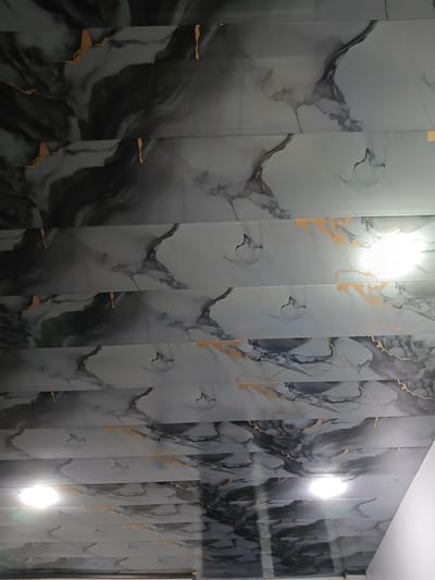 The image shows a ceiling with a marble-like pattern, featuring streaks of black, gray, and gold. The pattern is horizontal and divided into sections by visible lines, suggesting it is made of panels. There are two bright lights on the ceiling, one in the upper right section and another lower down. The overall impression is of an indoor space, possibly a room or hallway. The image does not contain any visual cues that would allow for the identification of its specific location within Xidirsho, Uzbekistan.