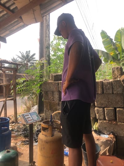 This image depicts an outdoor commercial scene in Alor, Nigeria, specifically a gas refilling or vending point.
A male individual, seen from the side facing right, stands centrally. He wears a purple short-sleeved shirt, black shorts, and a dark baseball cap, with his right hand on his hip and his head tilted downwards, observing the objects in front of him. To his left, a white digital weighing scale on a dark stand displays unreadable numbers. Directly in front of him and to the left of the scale is an upright, yellow liquid petroleum gas (LPG) cylinder with a valve and hose assembly attached to its top. Partially visible to the far left are a blue LPG cylinder and a green LPG cylinder on the ground.
The immediate ground is dirt, interspersed with some refuse, including plastic bottles and dark bags. In the background to the left, a wooden stall-like structure with shelving and a corrugated metal roof is visible, with a dirt road beyond it. Further back, dense green foliage and several palm trees are present. To the right of the individual, a stack of concrete blocks forms a wall, behind which more green vegetation is visible, including large-leafed plants. Overhead, a corrugated metal roof supported by wooden beams is partially visible, along with a concrete pillar. The scene is illuminated by bright daylight, under a clear or brightly overcast sky.