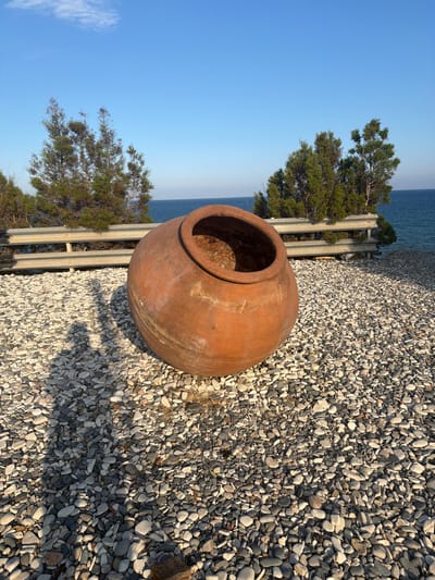 A large, reddish-brown terracotta pot rests on a bed of smooth, grey and white pebbles. The pot is round and wide, with a circular opening at the top. Its surface is textured and shows signs of age. In the background, a metal guardrail runs horizontally, separating the pebble area from a body of water, likely the sea, which is a deep blue. Behind the guardrail, several green, conical trees stand, their branches reaching upwards. The sky above is a clear, pale blue. The lighting suggests it is either early morning or late afternoon, casting long shadows from the pot onto the pebbles. The scene is located in Pervolia, Cyprus.
