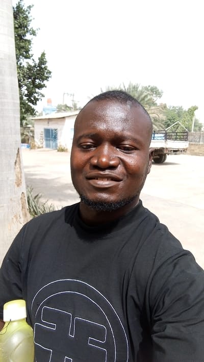An adult Black male is depicted in a brightly lit outdoor setting in Dakata, Nigeria. He is positioned in the foreground, facing the camera directly, and exhibits a slight smile, revealing teeth. He has a short, dark haircut and a short beard. The male wears a black short-sleeved t-shirt featuring a prominent white circular logo containing an 'F'-like symbol on the chest. In his left hand, partially visible in the lower-left corner, he holds a translucent yellow-green plastic bottle with a white cap. To the far left, a large, light-colored tree trunk is visible. The background includes a small, light-colored building with a blue door to the left, and a light-colored pickup truck with an empty bed positioned further right. Scattered green vegetation, including palm trees, is present in the mid-ground. The ground is a light-colored, paved or hard-packed surface, under a bright, clear sky.