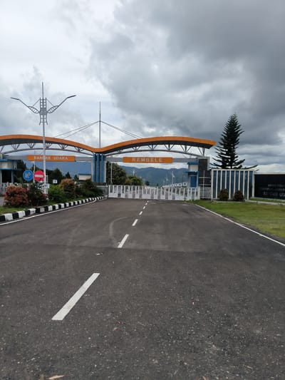 The image shows the entrance to Rembele Airport in Babussalam, Indonesia. The airport entrance features a large, modern archway with orange accents. The words "BANGLAR UDARA" and "REMBELE" are visible on signs under the archway. A paved road leads to a white gate, which is currently closed. The area is landscaped with trees and bushes, and there are mountains in the background. The sky is overcast with dark clouds, suggesting a cloudy or possibly rainy day. The road has white markings indicating lanes. A tall, slender evergreen tree stands to the right of the entrance. There are no people visible in the image.