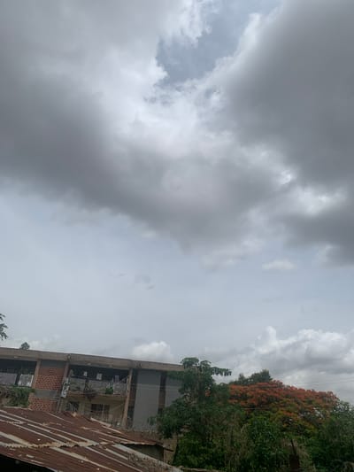 The image depicts an overcast sky with heavy, dark gray clouds dominating the upper portion. Below the clouds, the scene shows the rooftops of several structures. In the foreground, there is a rusty corrugated metal roof. Behind this, a multi-story building is visible, characterized by a brick facade on the left and a gray painted exterior on the right, with balconies featuring white railings. Various trees and foliage are present, including one with vibrant orange flowers to the right of the building. The overall impression is of a cloudy day in an urban or semi-urban environment, with elements of residential architecture and natural growth. The location in Kabong, Nigeria, is not directly evident from the visual information alone but is provided as context.