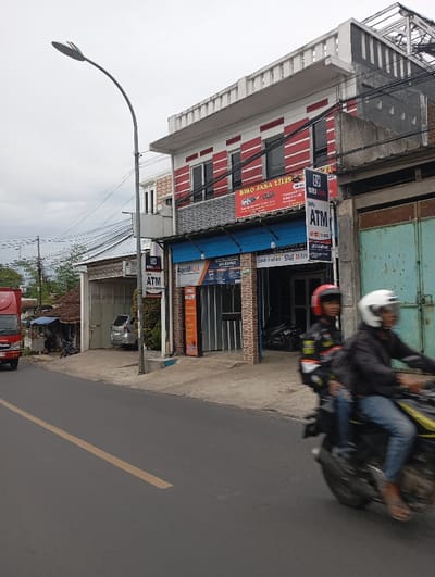 The image shows a street in Kecamatan Kutawaringin, Indonesia, with a two-story building on the right side of the frame. There is a sign on the building that reads "BIRO JASA Lilis," which is a local business. There is an ATM machine and a motorcycle parked in front of the building. A man is sitting on a motorcycle, wearing a helmet and looking ahead. In the background, there is a truck parked on the street. The sky is cloudy and the street is paved with asphalt.  There is a yellow dividing line on the road.  The image was taken on a street with power lines running overhead. The street is flanked by a sidewalk on the right side of the image.  There is a streetlight positioned above the sidewalk.  There is a small, white car parked on the left side of the frame. The image is shot from street level.