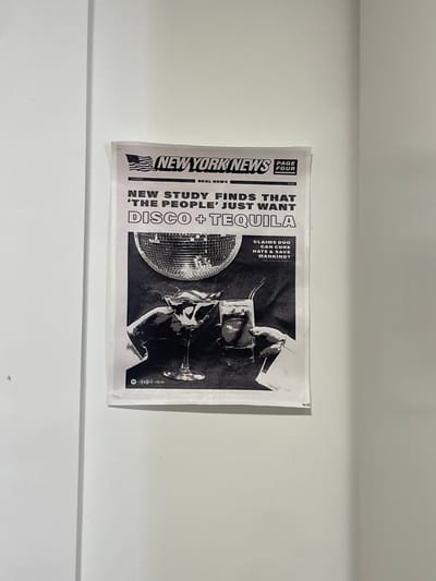 A black and white poster of a New York News article is adhered to a white wall. The headline reads, "NEW STUDY FINDS THAT 'THE PEOPLE' JUST WANT DISCO + TEQUILA" with an image of a disco ball, a martini glass, and a glass of tequila being toasted. The article claims that a music duo can cure hate and save mankind. The poster is located on a wall in an apartment in Segovia, Spain at coordinates 40.951, -4.119.