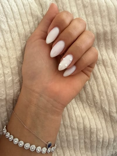 The image presents a close-up view of a person's left hand, adorned with a manicure and a bracelet, resting on a textured, light cream-colored fabric surface, possibly a ribbed blanket or towel.

The hand features almond-shaped nails painted in a solid white polish. The nail on the ring finger is distinctly embellished with three small, white, pearl-like beads arranged vertically.

A sophisticated silver-toned bracelet is worn on the wrist. It consists of interconnected circular segments, each meticulously set with multiple clear, sparkling gemstones, likely diamonds or crystals. A prominent design element is a teardrop-shaped dark blue gemstone, resembling a sapphire, encased in a silver setting, positioned near the top of the wrist. A delicate silver chain extends from this blue gemstone, curving across the back of the hand to connect with another part of the main bracelet, creating a subtle hand-chain or slave bracelet style.

The background fabric has a distinct horizontal ribbing pattern, adding to the soft texture of the scene. The lighting is soft and even, highlighting the details of the nails and the reflective quality of the bracelet's gemstones. No specific activity is taking place other than posing the hand to showcase the accessories. There is no visible text within the image. While the context suggests Cannes, France, no specific geographical indicators are present in the visual content.