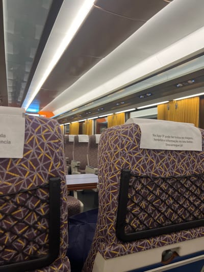 This image depicts the interior of a passenger train car, specifically identified as part of the Comboios de Portugal (CP) service, indicative of its operation within Portugal, including the city of Porto. The perspective is from a seated position, looking forward down the aisle. The train car is empty of passengers.
The seating features upholstery with a distinct purple and yellow geometric pattern. Each seat is equipped with a white paper headrest cover; the one in the foreground on the right displays Portuguese text advertising the "CP App" for travel information ("Na App CP pode ter todas as viagens, horários e informação no seu bolso. Descarregue já!"). A black mesh pocket is attached to the back of the foreground seat on the right. Further down the aisle, a small, light-colored table is visible between two rows of seats. A dark blue bag rests on the floor between seats in the midground.
The ceiling is modern, with multiple long, linear LED light strips providing illumination. Smaller blue indicator lights are visible along the overhead panel above the window line on the right side. Windows are visible beyond the seating, partially covered by amber-yellow vertical blinds or curtains. The overall environment appears clean and well-maintained.
