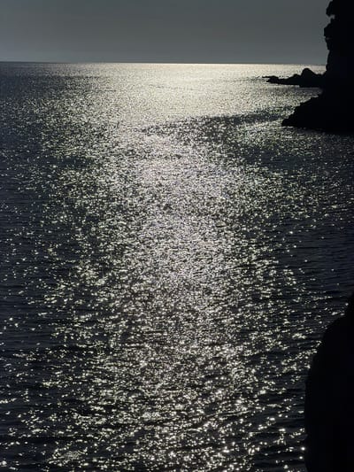 The image depicts a sun-drenched sea in Petrovac centar, Montenegro. The surface of the dark water is highly reflective, creating a shimmering effect from the sunlight. The horizon line is visible in the distance, separating the water from a dusky, overcast sky. To the right of the frame, a dark, silhouetted rocky coastline juts into the water. The overall scene is characterized by the interplay of light and shadow on the water's surface and the rugged coastal landscape. There are no people or distinct objects other than the natural elements visible.