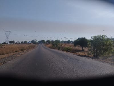 The image shows a paved road leading into the distance.  The road is located in Mararaban Tula, Nigeria (latitude: 9.756, longitude: 11.398). There are trees and brush lining the sides of the road. The road is straight and appears to be in good condition. The sky is blue and hazy with some wispy clouds.  There are electric power lines in the distance.  The road is empty of any vehicles or people.