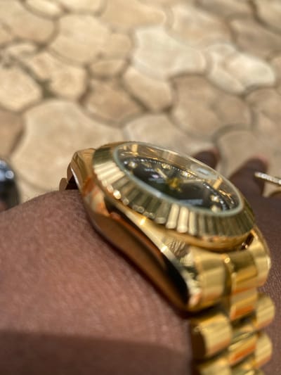 A close-up shot depicts a gold watch with a black face resting on dark skin. The watch has a fluted bezel and a gold band. A ring is visible on the finger. The background is a blurred image of textured, beige tiles. The city of Maigizo, Nigeria is not discernible from the image.