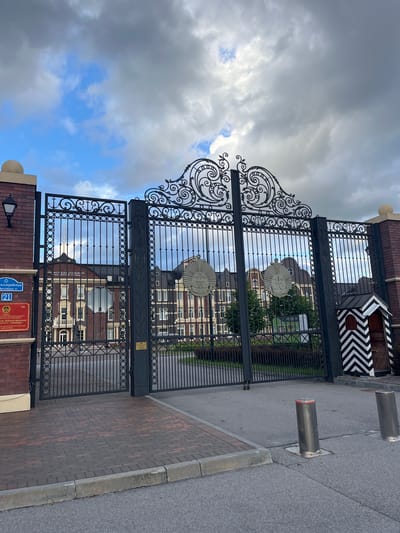 The image shows a large, ornate metal gate in front of a building. The gate has intricate scrolling metalwork at the top and a pattern of circles along the sides. Two circular emblems are visible on the gate, one on each of the two main sections. Beyond the gate, a multi-story brick building with many windows is visible. The building has a dark roof and a prominent central structure topped with a dome.

To the left of the gate, a brick pillar displays two signs. The upper sign is rectangular with blue and white text, and the number "21" is prominently displayed. Below it is a red rectangular sign with a coat of arms and Cyrillic text. A lamppost is mounted on this pillar.

To the right of the gate, a small, striped guardhouse with a black and white chevron pattern stands. The area in front of the gate is paved with red and brown bricks, bordered by a grey concrete curb. The street in front of the paved area is asphalt. Two silver bollards are visible on the asphalt, one of which has red lights on top.

The sky above is cloudy with patches of blue sky visible. The scene is likely a formal entrance to an institution or government building. This location is identified as the Kaliningrad Higher Naval School, also known as the Admiral Ushakov Naval Institute, located at 21 Komsomolskaya Street in Kaliningrad, Russia.