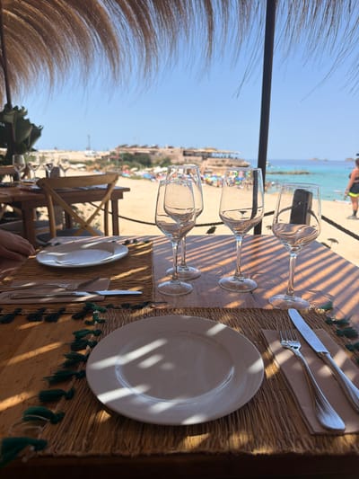 This image depicts a beachfront restaurant setting in Sant Josep de sa Talaia, Spain. The scene is set on a wooden table covered with straw placemats adorned with green tassels. On each placemat, a white plate is set, with cutlery (a fork and a knife) to the right. Four empty wine glasses are arranged on the table. In the background, the turquoise waters of the Mediterranean Sea are visible, along with a sandy beach populated by people and beach umbrellas. The sky is clear and blue. The restaurant appears to be open-air, with a thatched roof overhead casting striped shadows on the table. A person's hand is visible on the left side of the frame, suggesting someone is seated at the table.