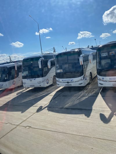 The image shows a row of white Велесстрой buses parked on a concrete surface under a blue sky with scattered clouds. The scene appears to be outdoors in Koskolovo, Russia. The buses are lined up closely, casting long shadows on the ground. Street lights are visible in the background.