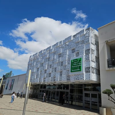 The image shows a modern building with a distinctive facade in Fez, Morocco. The building's exterior is covered in white panels featuring a perforated pattern and Arabic script design. A large green sign with Arabic text is prominently displayed. The building has glass windows and doors, allowing a glimpse of activity inside, with individuals entering or exiting. The surrounding environment includes a paved pedestrian area with streetlights, other buildings, and signage. The sky is blue with white clouds. People are walking along the sidewalk, suggesting a public space.