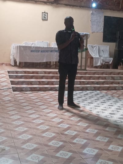 The image shows a man standing on a tiled floor in a room. The man is wearing a dark shirt and black pants. He is holding a microphone in his right hand and speaking into it. The man is standing in front of a table with a white tablecloth that reads "MAKUACHE MONTHLY MEETING". There are also two white chairs in the background. The floor is made of beige and white tiles with a floral design. The image was taken in Malava, Kenya, at a location with coordinates 0.53 degrees latitude and 34.813 degrees longitude. The image is taken at a low angle, looking up at the man. The lighting is bright and the room is clean and well-maintained. The man appears to be giving a speech or presentation. The image is likely taken during a community meeting or gathering.