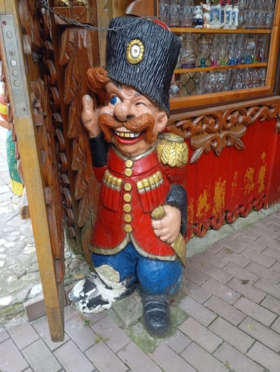 An outdoor, eye-level, full shot captures a wooden sculpture of a Cossack figure. The sculpture depicts a man with a large red jacket adorned with gold buttons, blue pants, and black boots. He has a wide, toothy grin, blue eyes, and a thick, reddish-brown mustache and beard. He wears a tall, black, fur-trimmed hat with a gold emblem. The figure is standing on a cobblestone pavement. Behind the sculpture, a red wooden structure with intricate carvings is visible, and a wooden display cabinet filled with glassware is mounted on it. To the left of the sculpture, a wooden post with a metal panel containing several buttons is attached to a chain. The general location is identified as Goryachiy Klyuch, Russia.