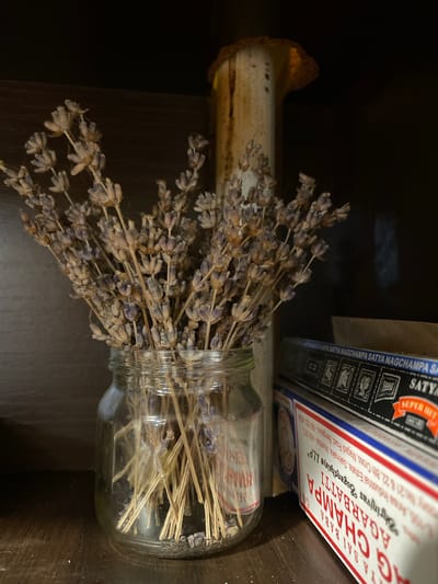 A close-up shot shows dried lavender in a glass jar on a wooden shelf. Behind the lavender, a rustic, aged candlestick or pillar stands, its top adorned with a burnt orange hue. To the right of the lavender, several boxes of incense sticks are stacked. The packaging of one box, in white with red and blue accents, displays the words "SAI BABA AGARBATTI". The overall setting appears to be indoors, possibly in a home or shop, with a dark wooden background. The lighting is dim, casting shadows and highlighting the textures of the dried lavender and incense boxes. There are no people or animals present in the image. The location within Sevastopol, Ukraine, cannot be determined from the visual information provided.