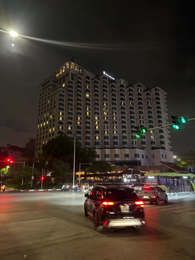 The image shows a night scene at an intersection in Hanoi, Vietnam, in front of the Pan Pacific hotel. The hotel is a tall, multi-story building with many illuminated windows. The intersection is busy with traffic, including cars, motorcycles, and a green bus. Traffic lights are visible, with green lights illuminated. The street is wet, reflecting the lights from the vehicles and buildings. The sky is dark with some clouds, and there is a bright light source, possibly the moon or a street lamp, with a lens flare.