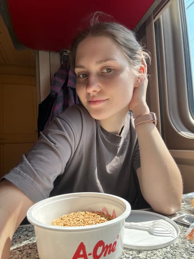 A young Caucasian woman with light brown hair pulled back is seated on a train, likely in Chudovo, Russia, judging by the context of the location. She is looking at the camera with a slight smile and has her left hand resting on her head. She is wearing a plain grey t-shirt and a light pink fitness tracker on her right wrist. In front of her, on a speckled surface that appears to be a table, is a white cup of instant noodles. A white plastic fork and plate are also visible. The background shows the interior of a train carriage, with wooden paneling, a red upholstered ceiling, and a window with brown trim. A plaid shirt is hanging on a hook behind her. The lighting suggests it is daytime, with sunlight streaming in from the window.