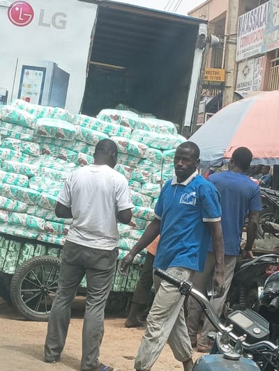 The scene depicts a bustling street in Fagge C, Nigeria, with a focus on commercial activity and the movement of goods. In the center, a white cargo truck, bearing an "LG" logo on its side, is parked with its rear door open, revealing a substantial quantity of stacked packages. These packages are large, rectangular, and wrapped in plastic with a recurring light blue/green and brown pattern, suggesting they contain items like diapers or sanitary pads. The packages are stacked high on what appears to be a wheeled, wooden handcart or trolley positioned directly behind the truck.

Three adult males are prominently featured. In the immediate foreground, slightly to the right, a man with dark hair and a light mustache is seen from the waist up, walking towards the right side of the frame. He wears a blue polo shirt with a white collar and sleeve cuffs, featuring a white logo of an open book and a quill on the left chest. His light-colored trousers have "OUR" visible on the upper left leg, and he is barefoot. His left hand is near the handlebars of a blue motorcycle, which is partially visible in the lower right foreground.

Behind this man and to the left, another man is seen from the back, wearing a white t-shirt and dark grey trousers, with blue flip-flops. He stands facing the stacked packages, appearing to be observing or handling them. Further to the right and slightly behind, a third man is partially visible from the back, wearing a plain dark blue t-shirt and light-colored trousers, also appearing barefoot.

In the background, street activity continues. Buildings with commercial signs are visible, including one prominently displaying "MAYASSALA COMMUNICATION," "No. 62, BABA NADEGU'S HOUSE," and a phone number. A yellow sign partially reads "[AROTH] ING TO PICK ING TO DROP." An open, orange and grey striped umbrella provides shade in the mid-background. The ground is unpaved, appearing as compacted dirt or sand. Several other individuals and motorcycles are present in the background, indicative of a busy urban environment.