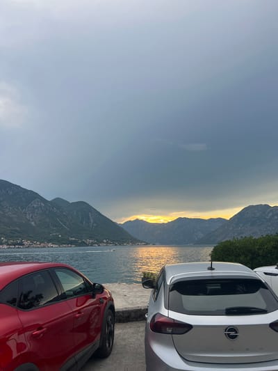 A scene of a bay in Dobrota, Montenegro is captured during a cloudy sunset. The foreground features the rear ends of a red car and a silver Opel Corsa parked on a concrete surface. Beyond the cars, a body of water stretches out, reflecting the warm hues of the setting sun. On the far side of the bay, a chain of lush green mountains dominates the landscape, with a small town nestled along their base. A single white boat is visible on the water, moving from left to right, leaving a white wake. The sky above is filled with dramatic, dark clouds, interspersed with patches of brighter, orange light where the sun is setting behind the mountains.