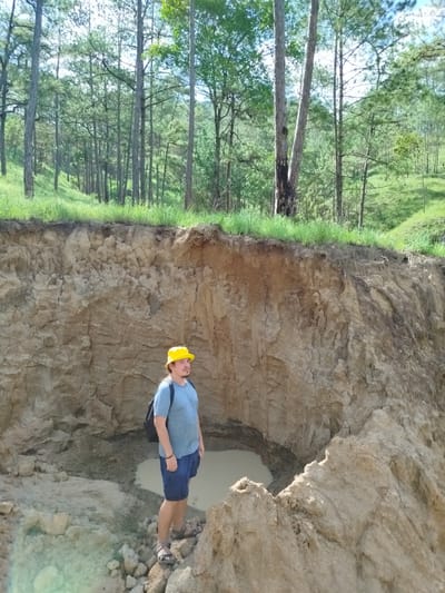The image captures an outdoor scene in Tuý Sơn, Vietnam, featuring one adult male subject situated within a large, excavated or naturally formed pit. The pit's exposed walls, composed of light brown, consolidated soil and rock, rise substantially higher than the man, displaying distinct textural variations, cracks, and strata. At the bottom of the pit, a pool of opaque, light brown water is present alongside loose soil and scattered rocks, on which the man stands.
The man, appearing to be of average build with light skin, is wearing a light blue short-sleeved t-shirt, dark blue shorts, and grey open-toed sandals. He has light brown hair, visible beneath a bright yellow bucket hat, and a moustache. A dark-colored backpack is worn on his left shoulder. He is oriented slightly to his right (viewer's left), with his head tilted upward and to his right, looking towards the top edge of the pit and the background.
Above the pit's rim, a strip of green grass marks the transition to an upward-sloping terrain densely covered by numerous tall, slender trees, primarily conifers, with green foliage. The sky, partially visible through the tree canopy, appears bright and clear, indicating a sunny day with strong natural illumination.