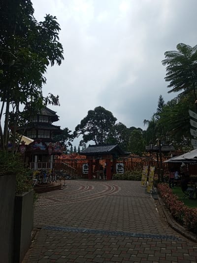 The scene depicts an outdoor complex, located in Tân Kỳ Commune, Vietnam, under a uniformly overcast sky. In the left midground, a multi-tiered structure, consistent with traditional Japanese architecture featuring dark tiled roofs and white facades, stands partially visible behind foreground trees. A small carousel ride with painted horses is positioned below this structure. A dark red, traditional Japanese gate, resembling a torii, with a tiled roof, marks an entrance in the center midground. A paved pathway of interlocking reddish-brown and gray bricks, detailed with concentric curved patterns, leads towards this gate. Multiple individuals are discernible near and beyond the gate structure, appearing to transit the area. To the right, an outdoor dining or seating area is present under a dark umbrella, where several individuals are seated or standing. A multi-directional signpost and two vertical yellow promotional banners are also visible on the right side of the pathway, surrounded by manicured shrubs. Varied trees, including broadleaf and palm species, frame the complex, which features additional structures and orange-colored fencing beyond the central gate.